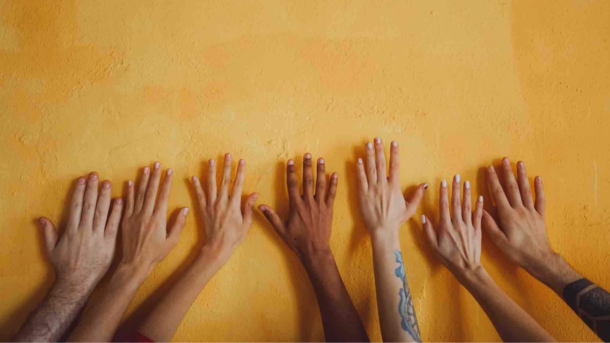 Image of variety of arms and hands together on a yellow wall.